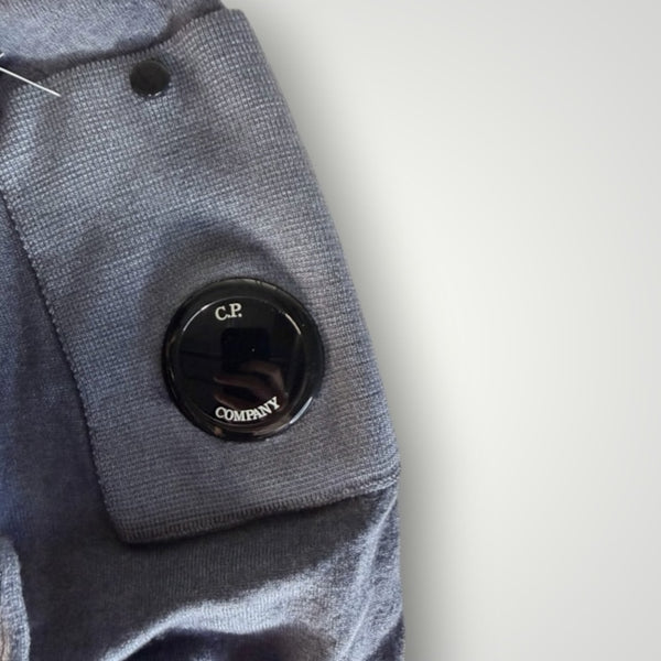 Gros plan sur la manche grise d'un pull Secondstore C.P. Company avec sa poche utilitaire et la lentille emblématique