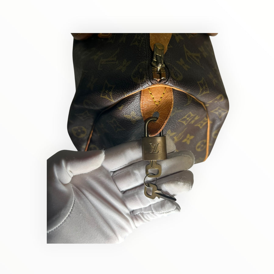 Main gantée de blanc tenant le cadenas doré d'un sac Secondstore Louis Vuitton Speedy en toile Monogram marron
